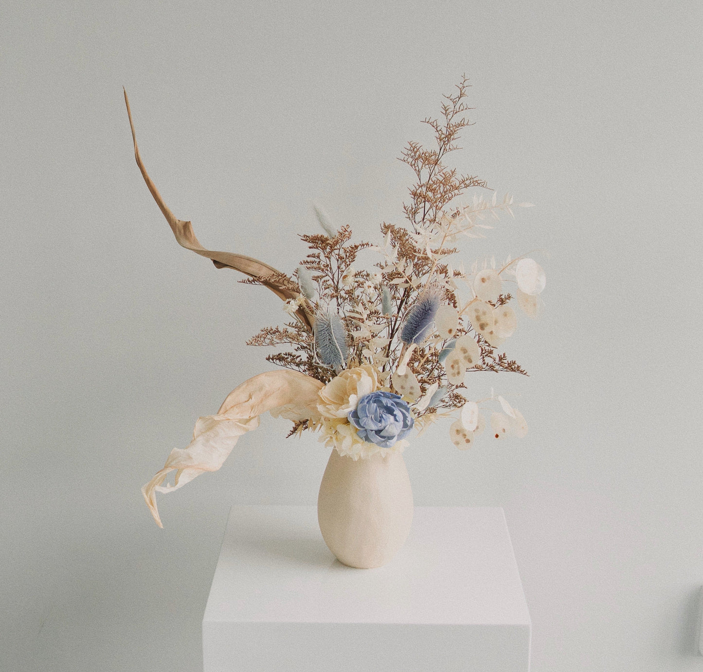 A ceramic vase filled with natural dried florals and a blue flower, arranged on a neutral background.
