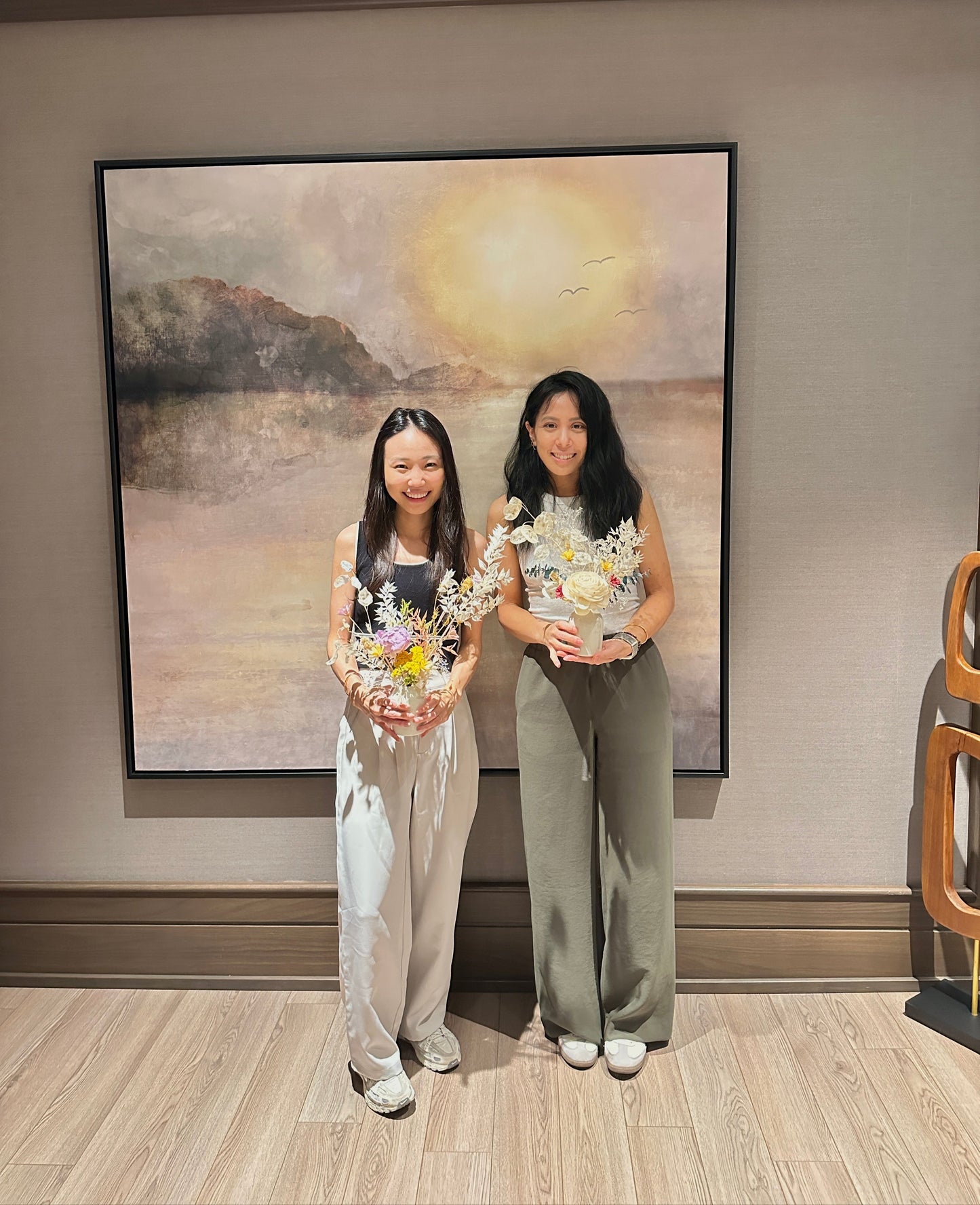 Two women standing in a room with flower arrangements in their hands in front of large painting of a landscape on the wall.