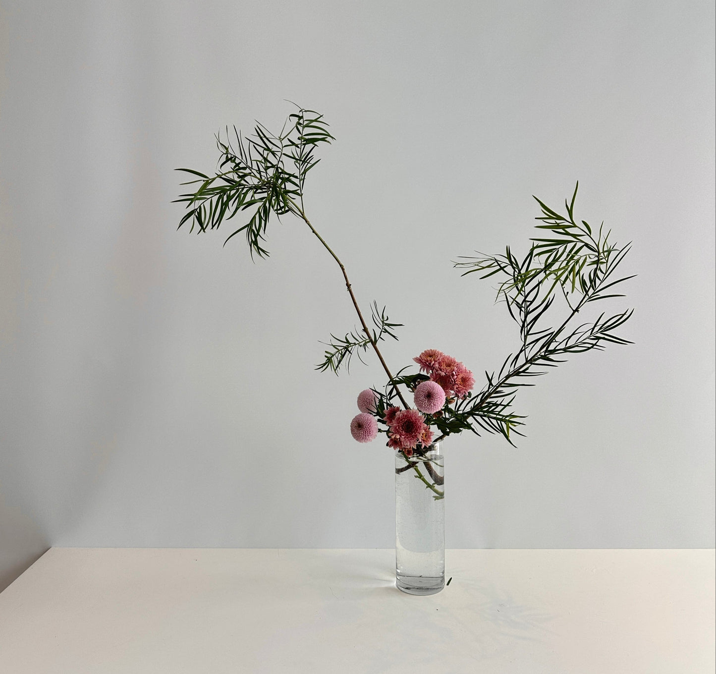 Glass vase with pink flowers and green leaves on a white table against a light gray background.