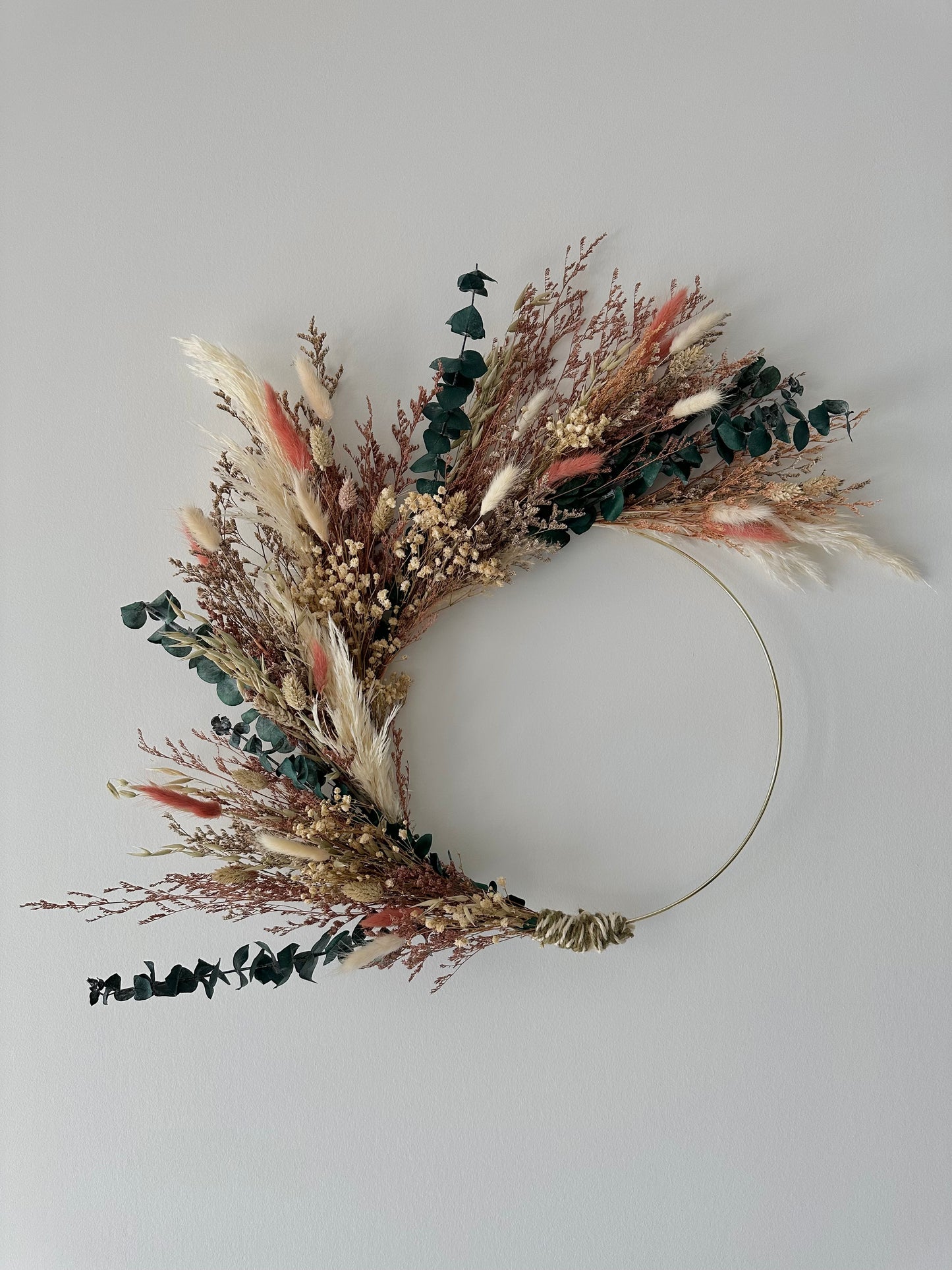 A dried floral wreath arrangement with a gold-colored hoop, featuring a variety of beige, brown, and multicolored flowers and foliage.