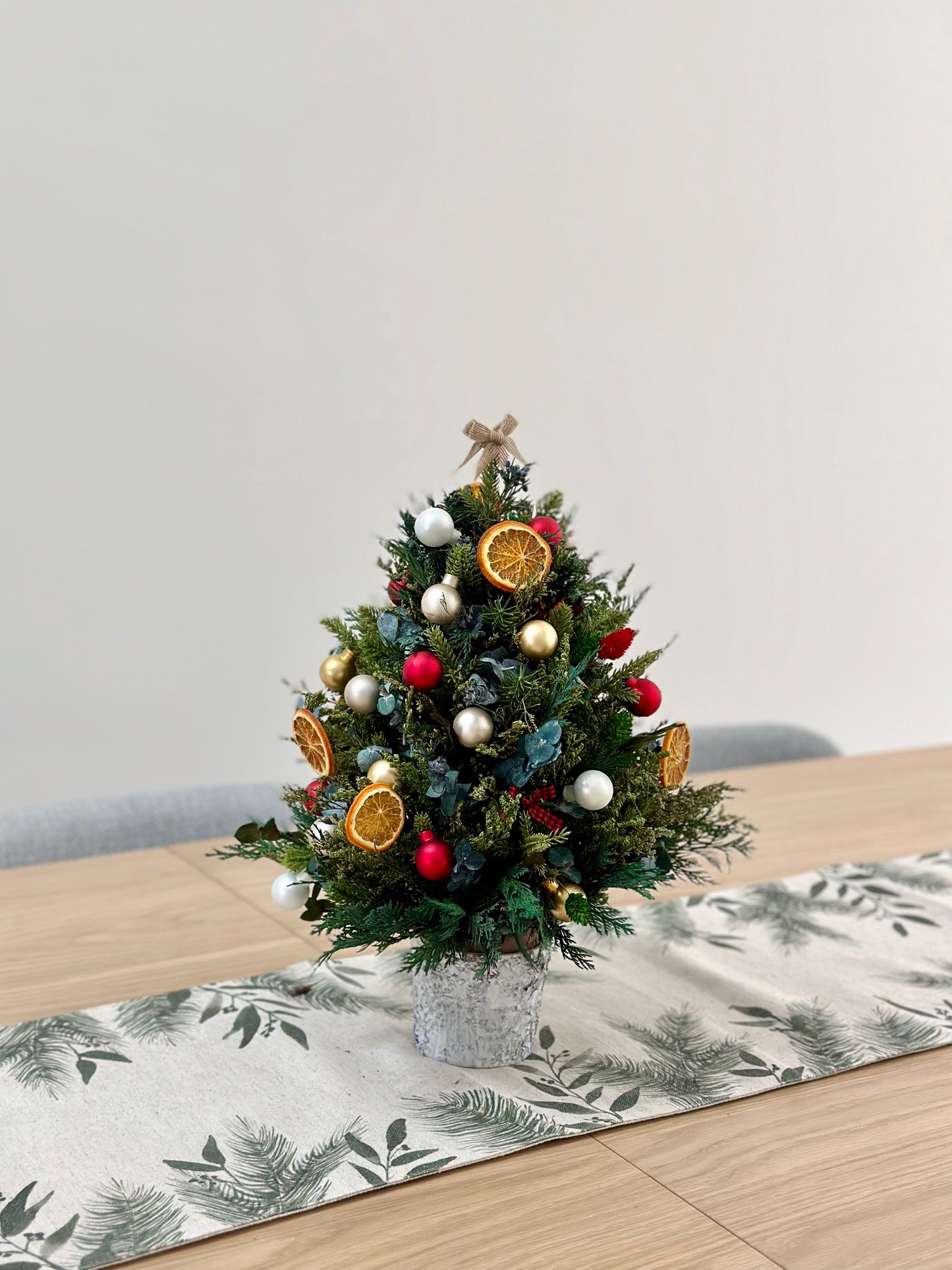 A mini Christmas tree made of dried flowers, featuring green branches and decorated with gold, red, and yellow ornaments, placed on a table with a patterned tablecloth.
