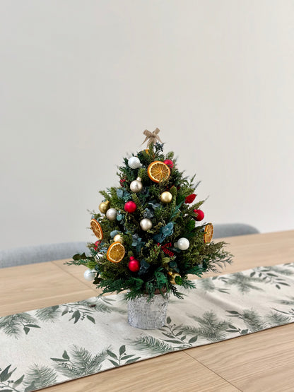 A mini Christmas tree made of dried flowers, featuring green branches and decorated with gold, red, and yellow ornaments, placed on a table with a patterned tablecloth.