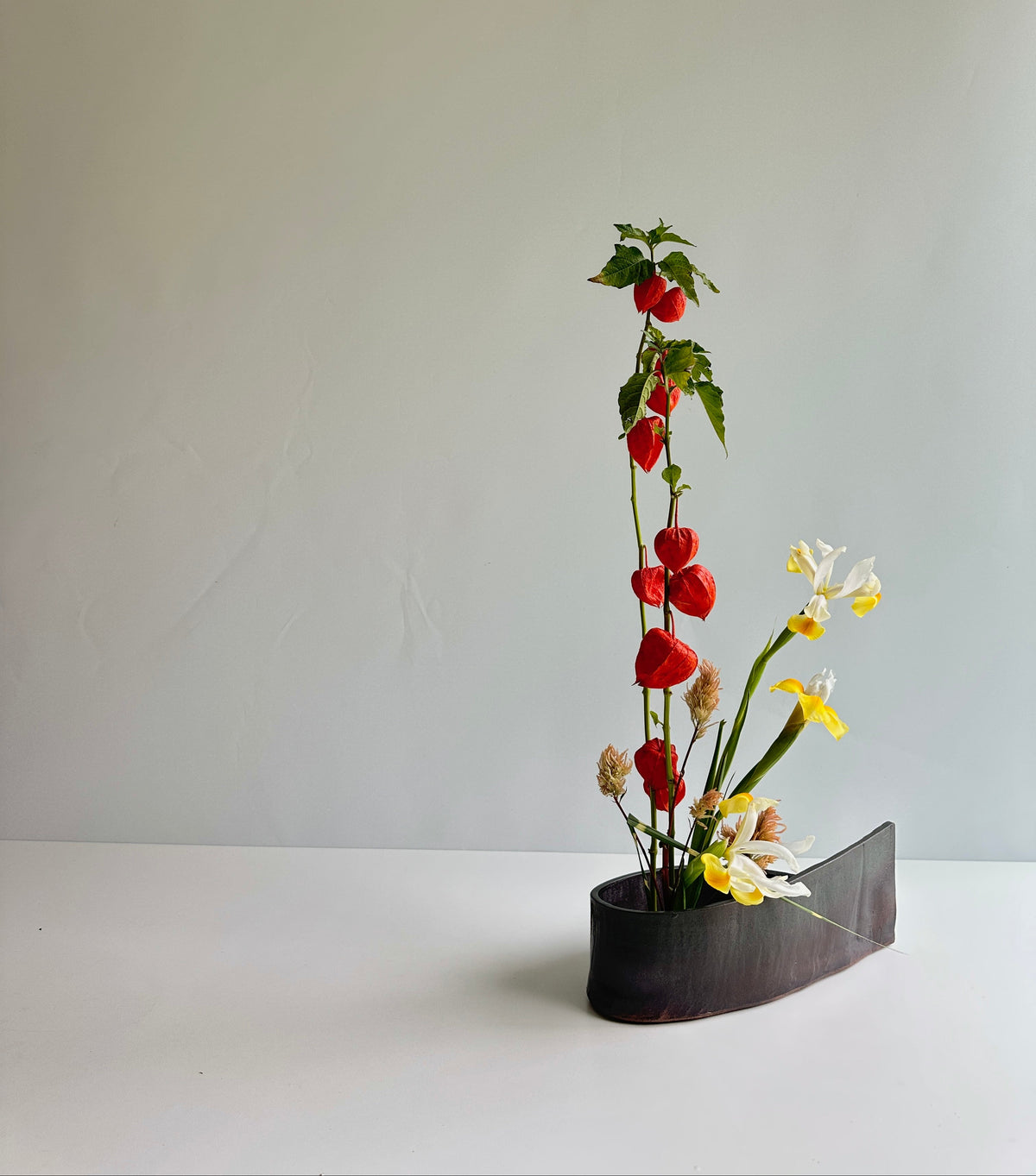 A minimalist floral arrangement featuring red berries on a tall stem, white and yellow flowers, and green leaves in a dark, sculptural ceramic vase, set against a plain light gray background.