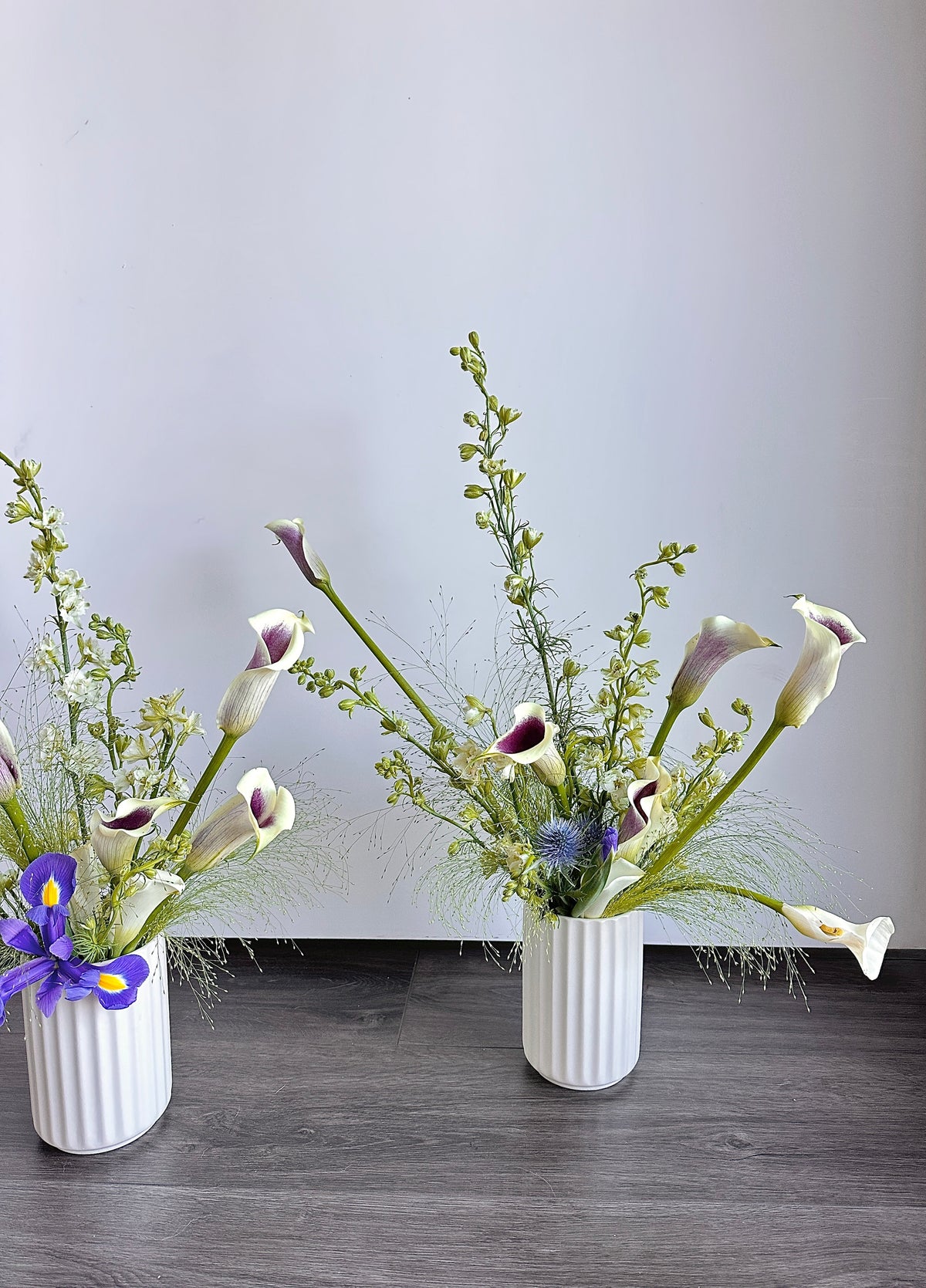 Two white ribbed ceramic vases filled with calla lilies and green filler stems, displayed on a dark wood floor against a plain light gray wall.