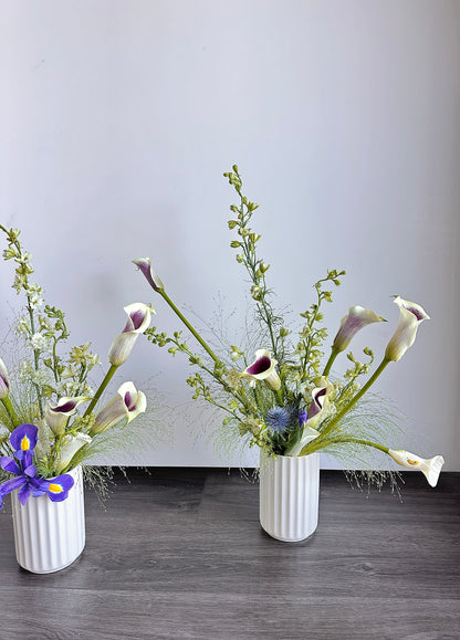 Two white ribbed ceramic vases filled with calla lilies and green filler stems, displayed on a dark wood floor against a plain light gray wall.