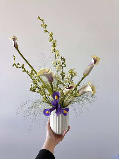 A hand holds a white ribbed ceramic vase filled with a floral arrangement featuring cream calla lilies, purple irises, and green foliage against a plain light gray background.