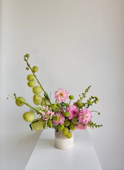 A floral arrangement featuring pink flowers and green seed pods in a small white ceramic vase, set on a white table against a light gray wall.
