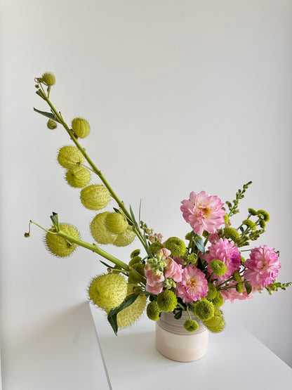 A floral arrangement featuring pink dahlia-like flowers and textured pale green seed pods in a small white ceramic vase, set against a clean white background.