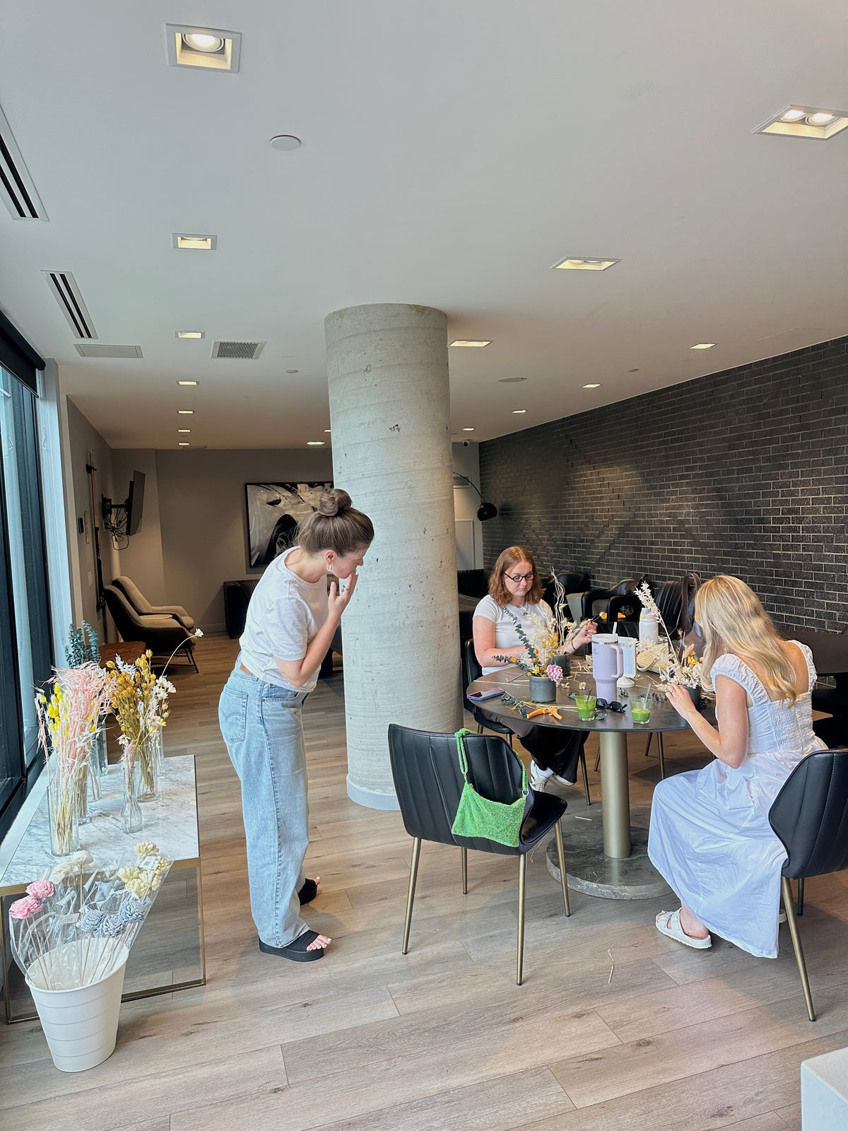 A woman in a white t-shirt and light blue jeans stands in a modern interior space, looking down with a sad expression. In the background, two women are seated at a round table with dried flowers in a contemporary room with a concrete column, brick wall, and wood floors.