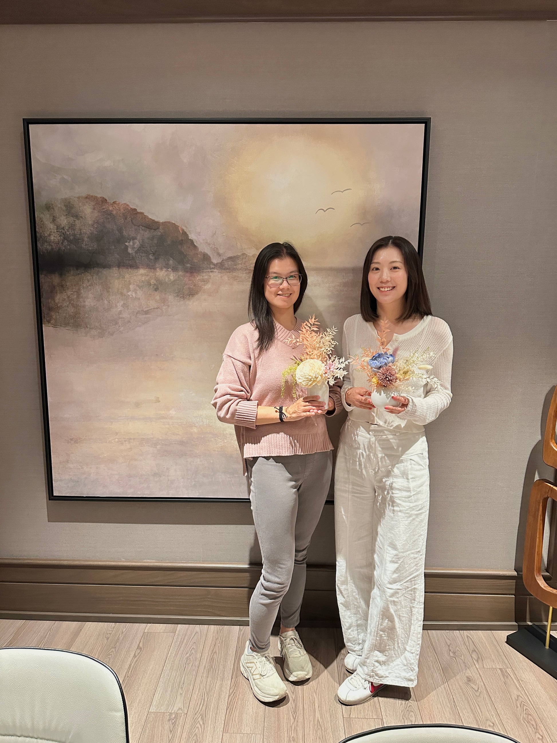 Two women standing with flower arrangements in front of a large painting of a landscape on the wall.
