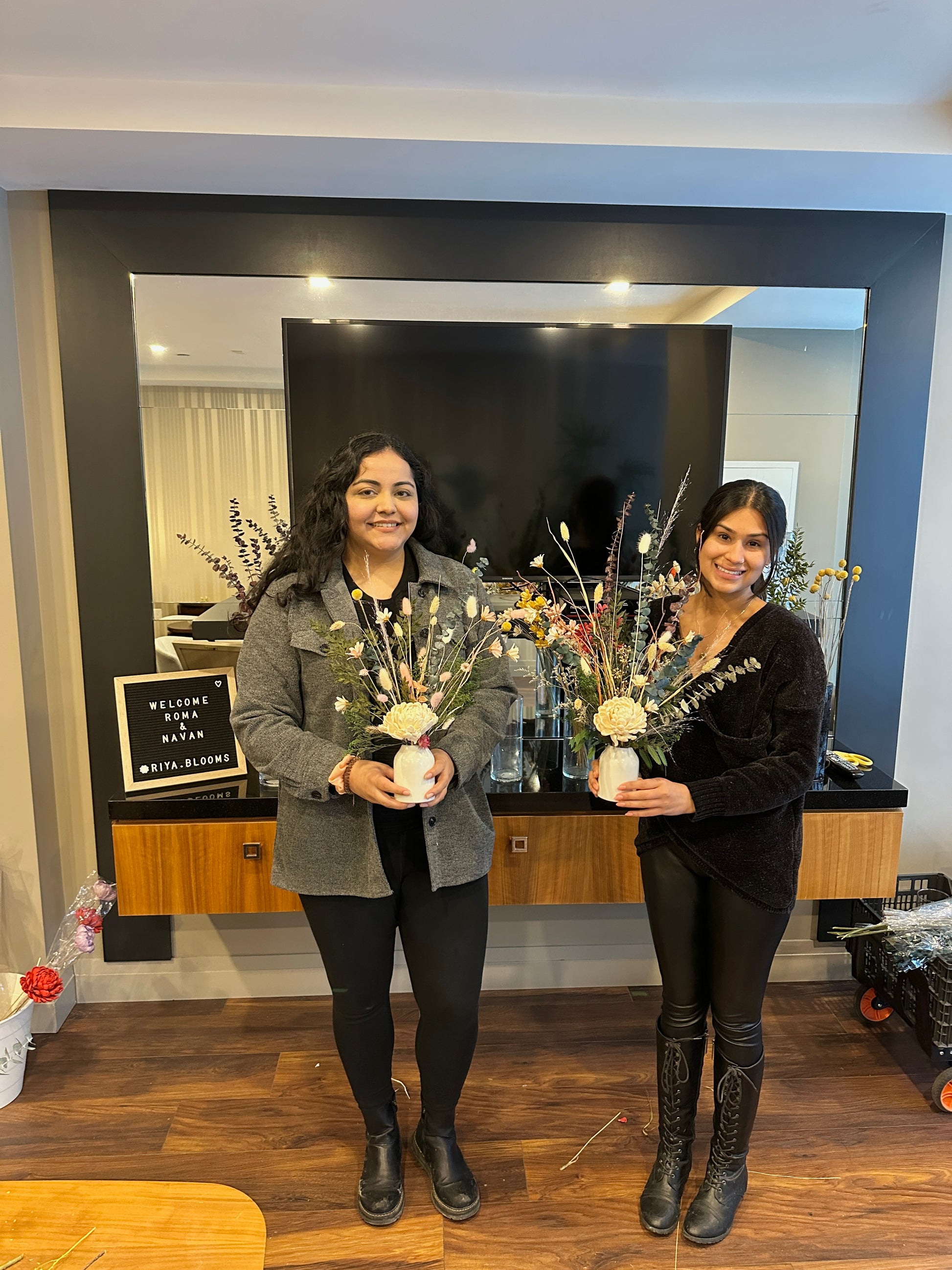 Two women holding flower arrangements in a room with a black wall and wooden floor.