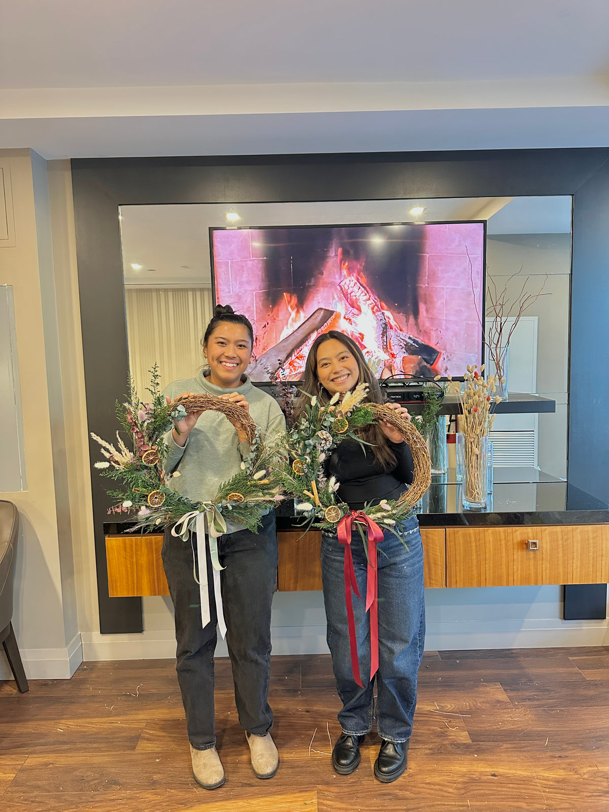 Two people holding decorative wreaths in a living room with a TV with fireplace in the background.