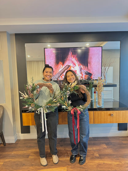 Two people holding decorative wreaths in a living room with a TV with fireplace in the background.
