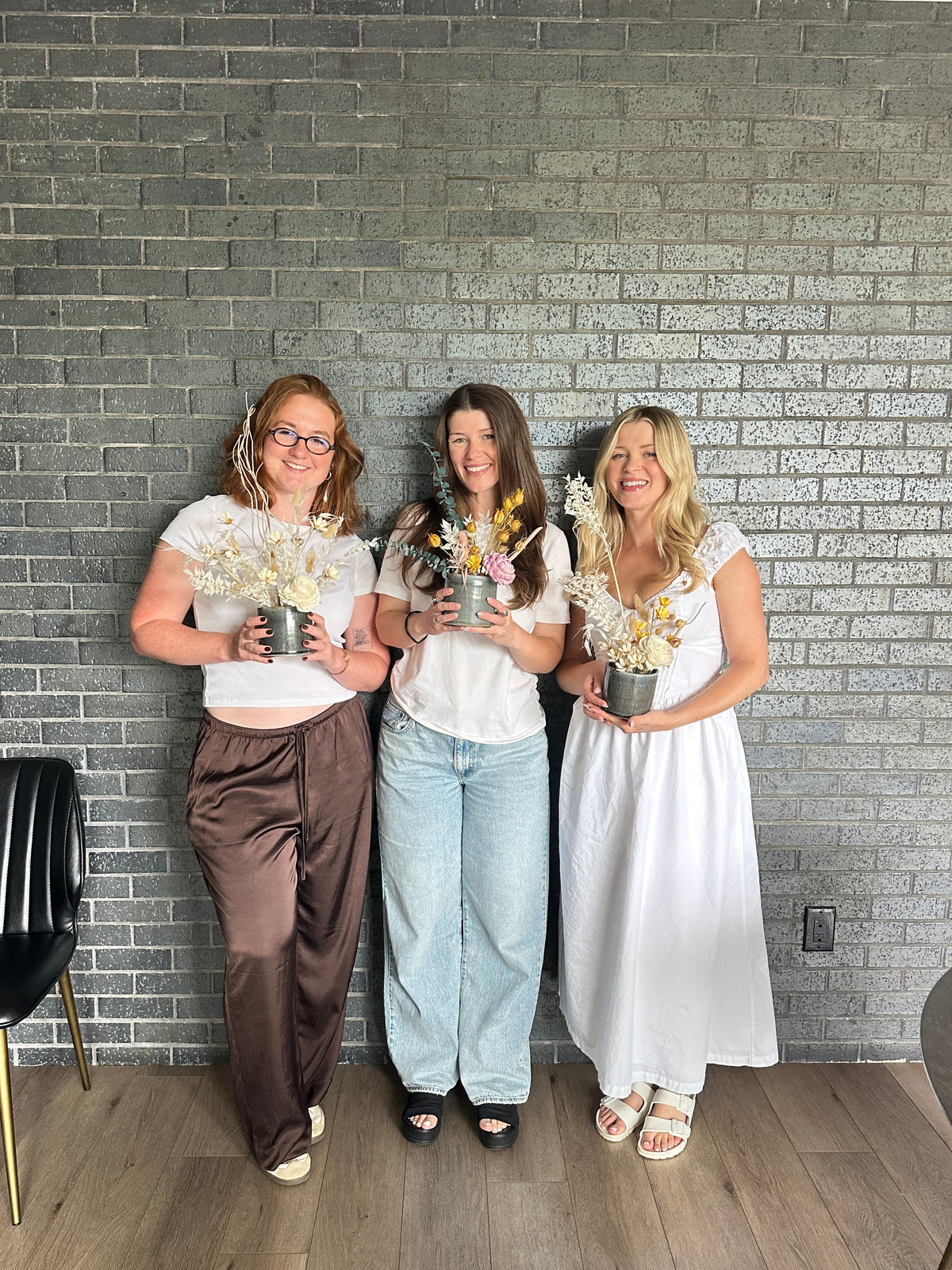 Three women standing together in front of a gray brick wall, each holding a small potted plant with dried flowers in a gray pot.