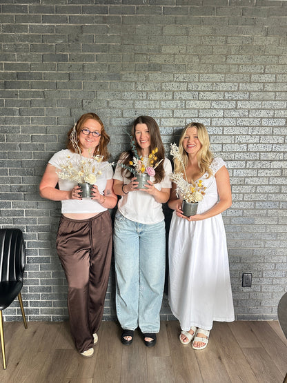 Three women standing together in front of a gray brick wall, each holding a small potted plant with dried flowers in a gray pot.