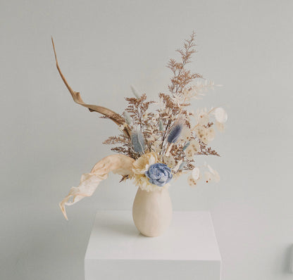 A ceramic vase filled with natural dried florals and a blue flower, arranged on a neutral background.