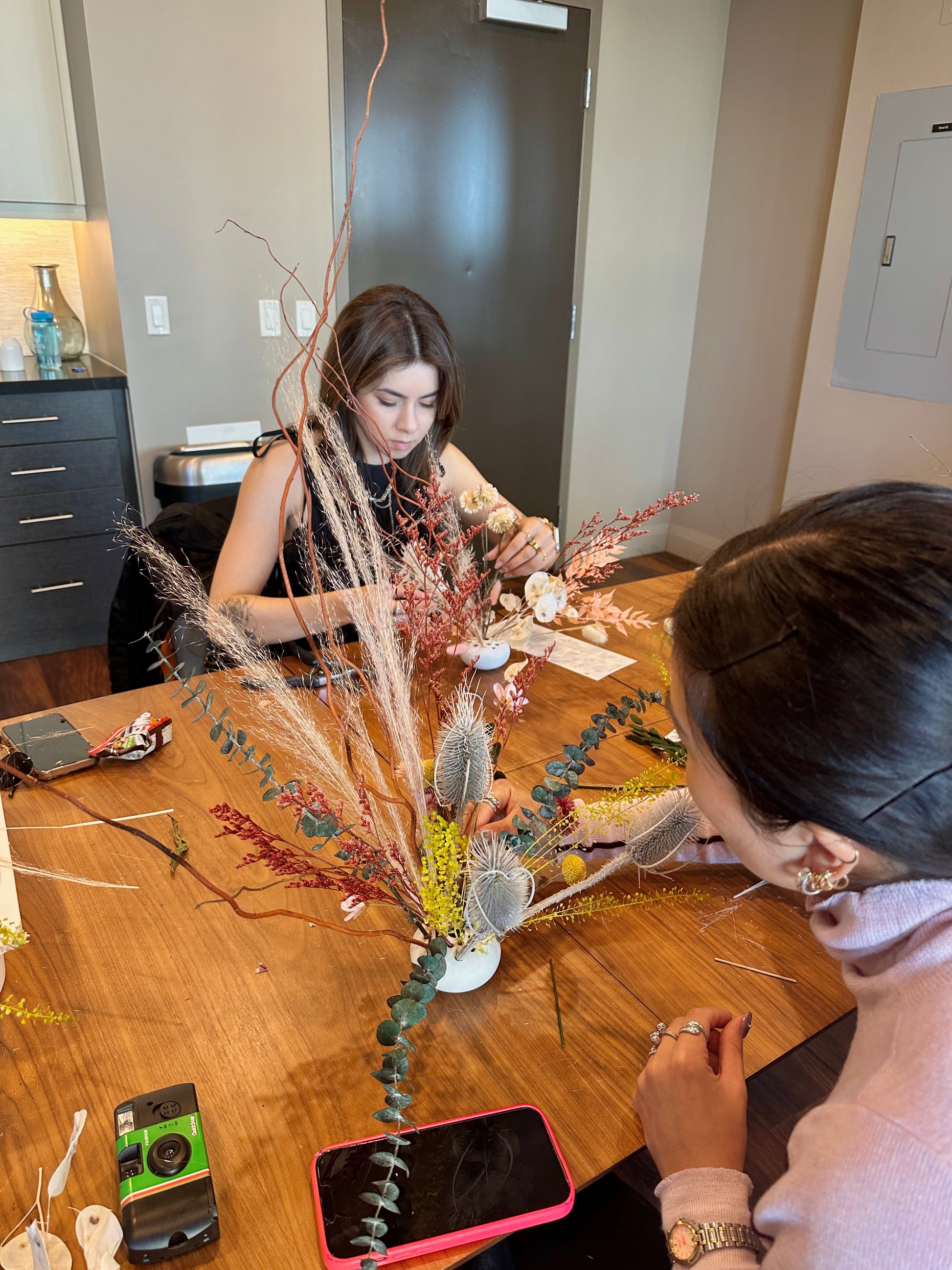 Two people working on a floral arrangement at a table in a room.