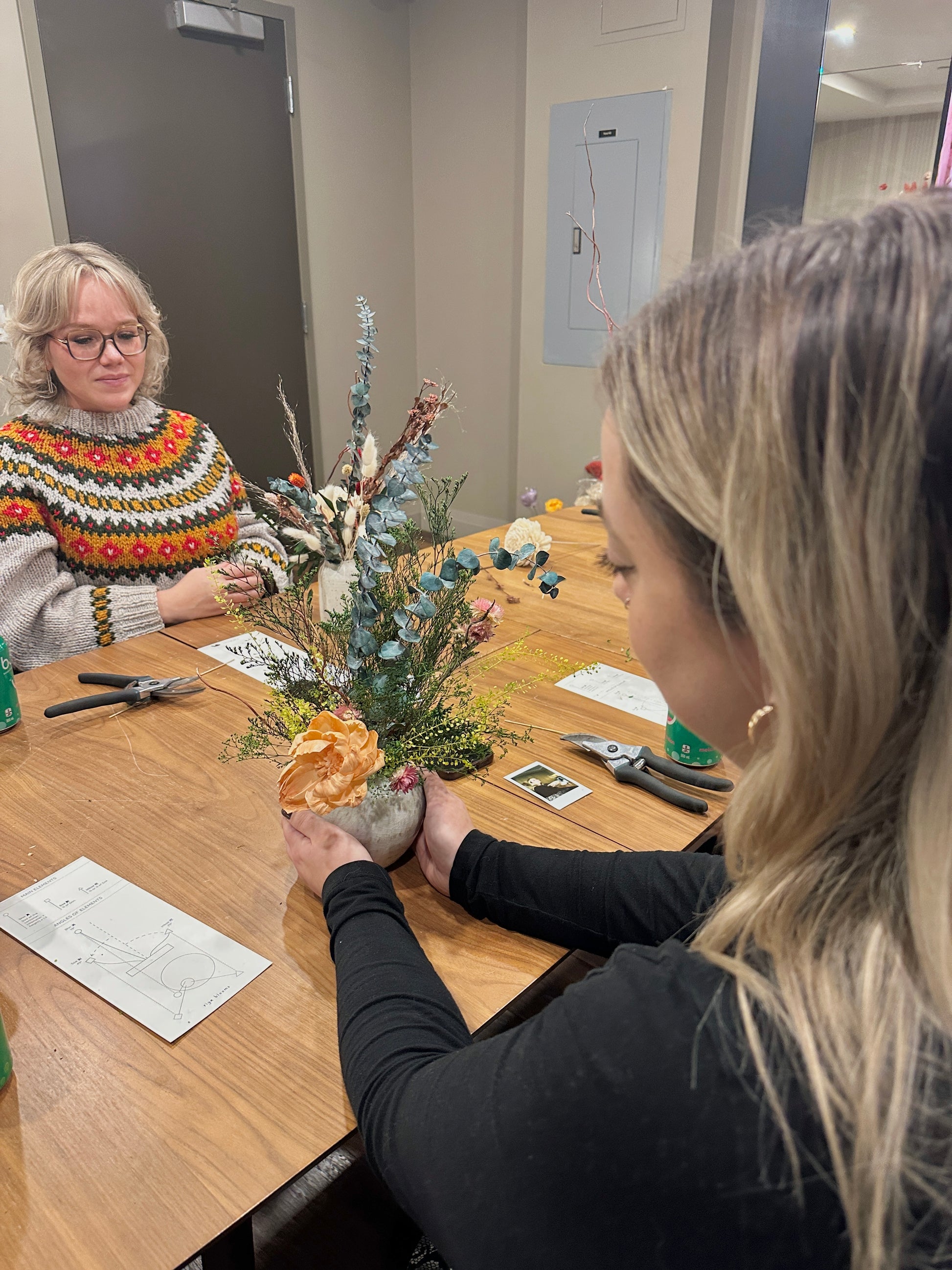 Two women working on a craft project at a table with floral materials and tools.