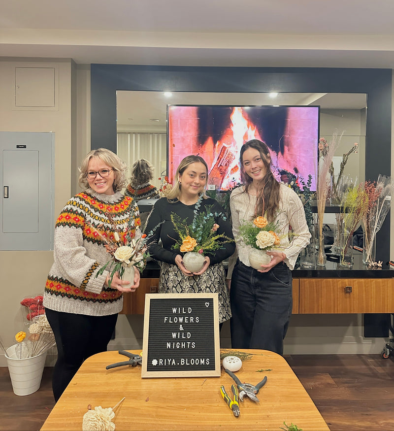 Three women holding floral arrangements in a room with a TV screen displaying a fireplace.