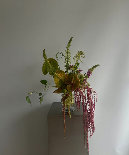 A styled floral arrangement featuring olive green anthuriums, burgundy flowers, and cascading pink amaryllis in a clear glass vase, displayed on a minimalist gray pedestal against a neutral light gray wall.
