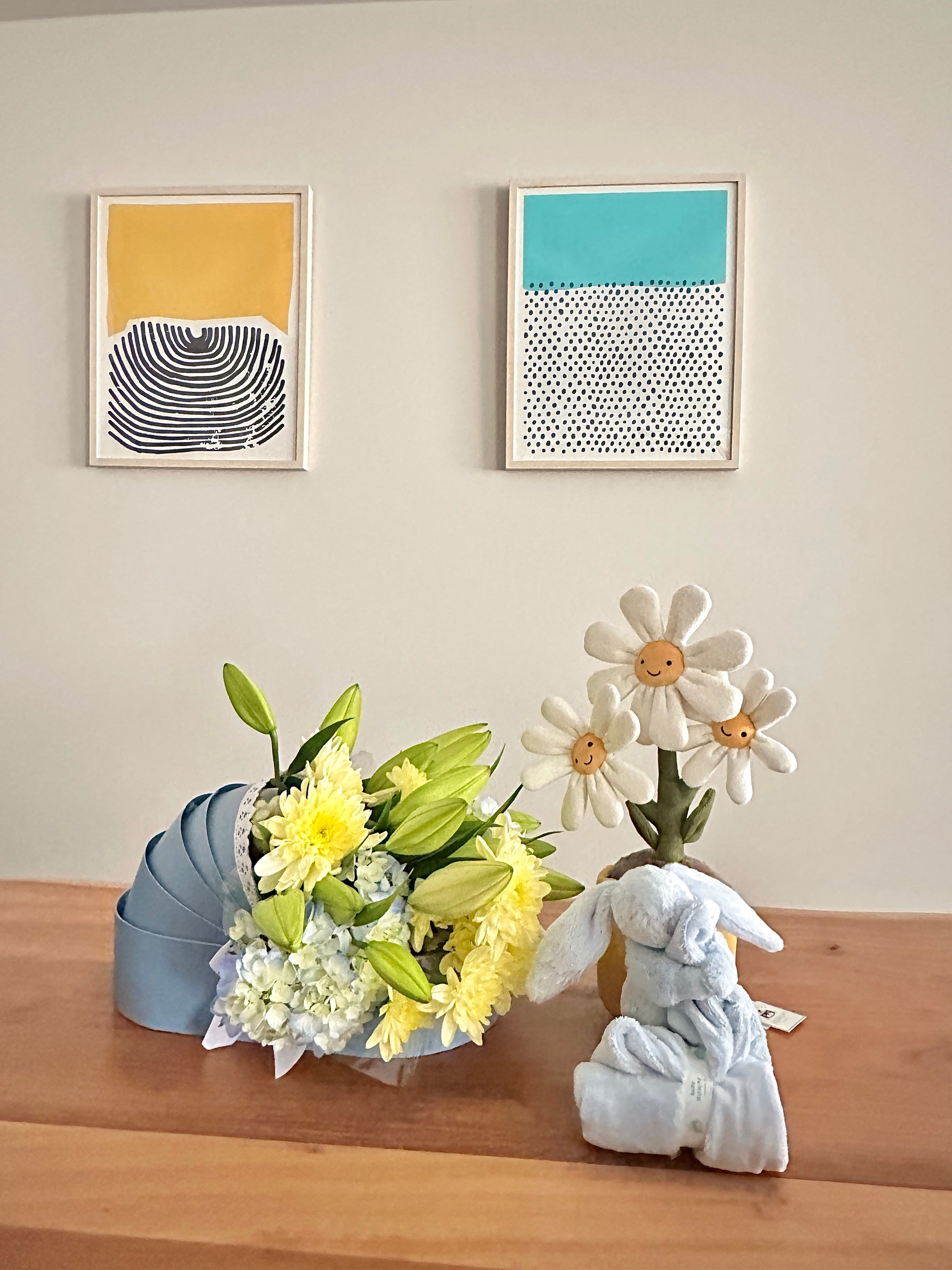 Bouquet of flowers, wrapped gift, and decorative bunny on a wooden surface with framed artwork in the background.