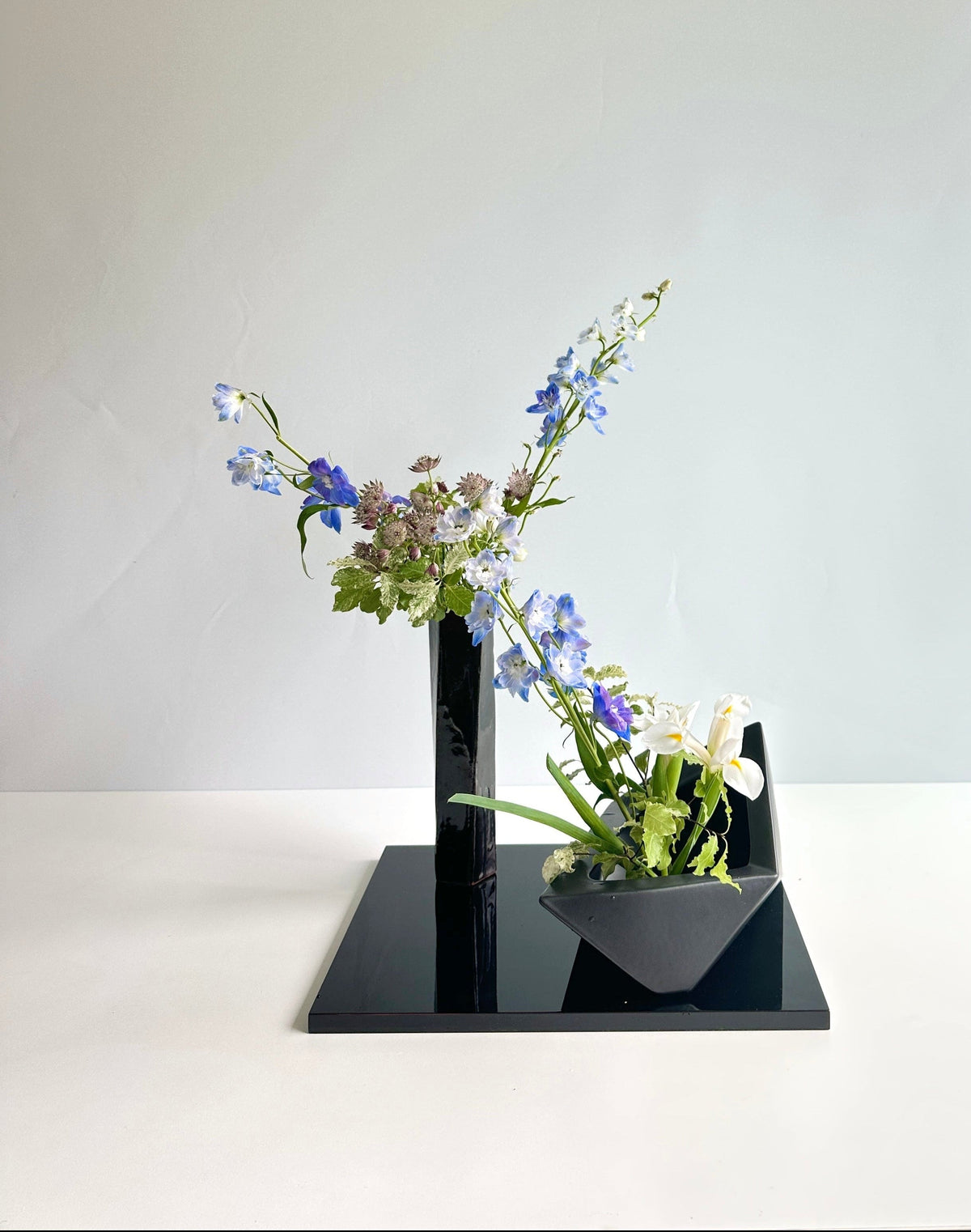 A minimalist black ceramic flower arrangement stand on a white surface against a light gray background, holding blue, purple, white, and green flowers.
