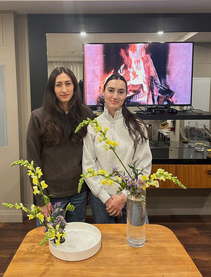 Two women standing with ikebana arrangements in a living room with a television and wooden table.