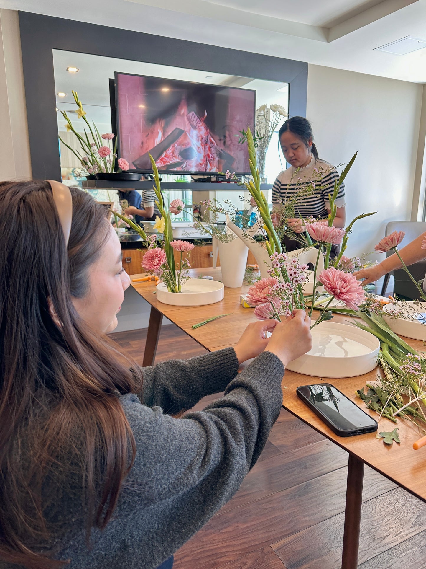 Two women arranging flowers in a home setting with a television in the background.