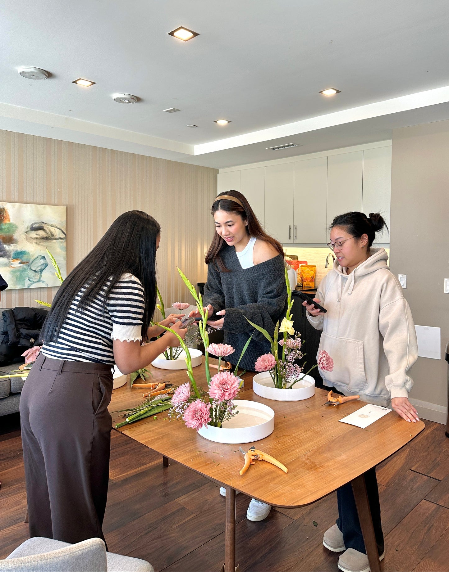 Three women in a kitchen setting, one is arranging flowers on a table.