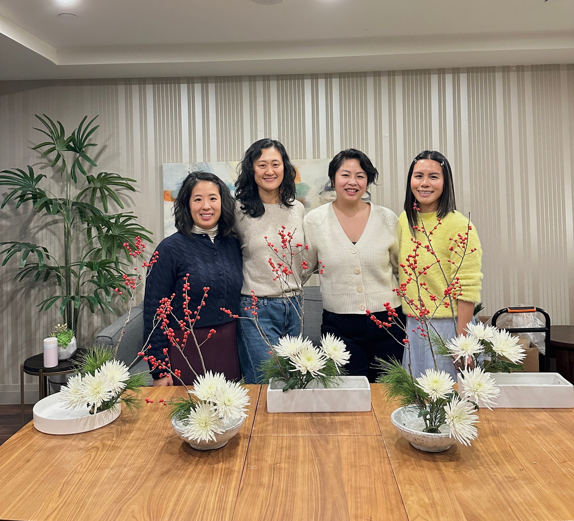 Four women posing together in a room with floral arrangements on a table.