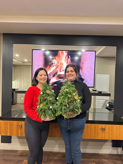 Two people standing indoors holding small decorated Christmas wreaths, with a large screen and wooden furniture in the background.
