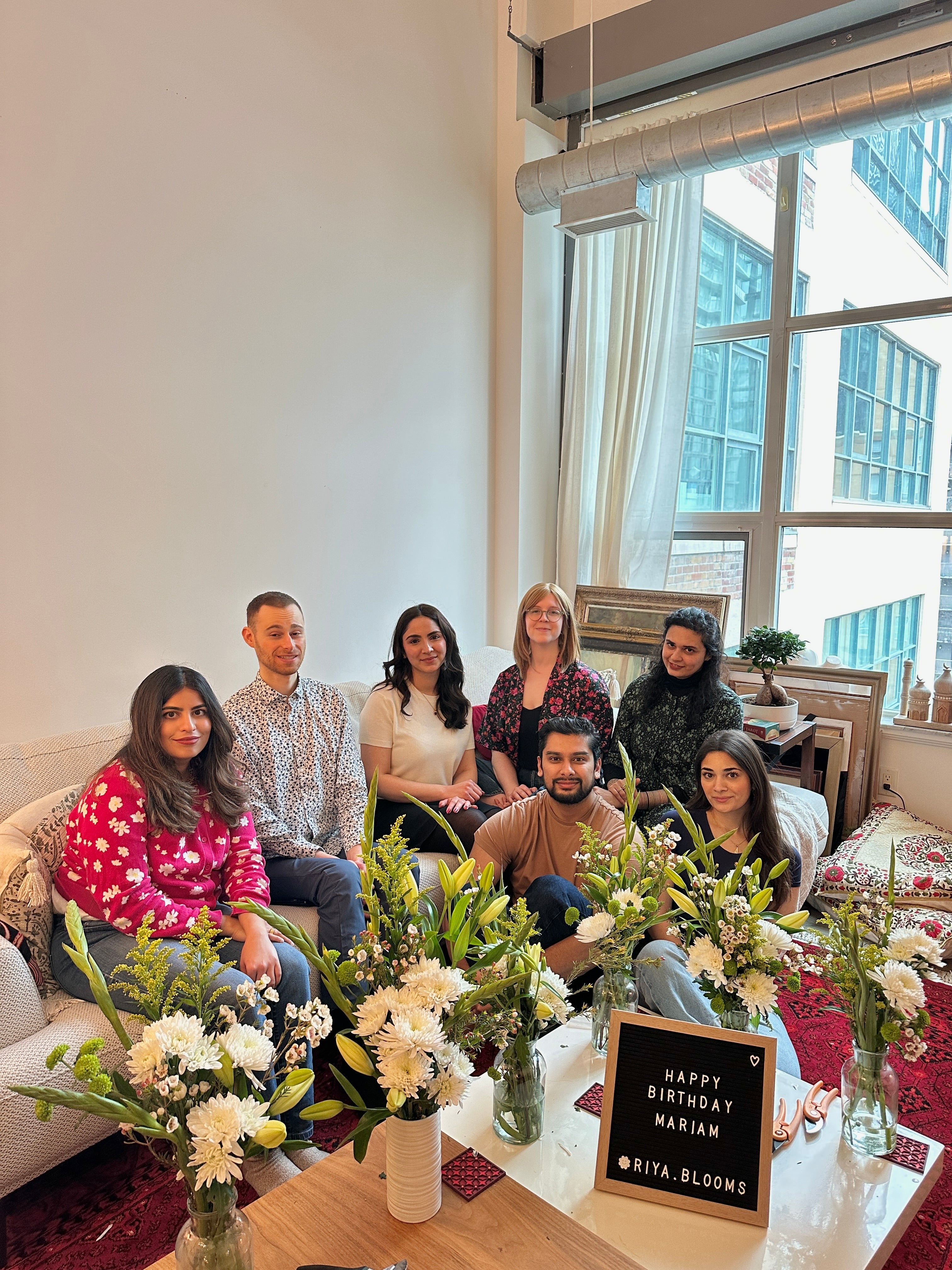 Group of people sitting together with floral arrangements and a 'Happy Birthday' sign in a modern living room.
