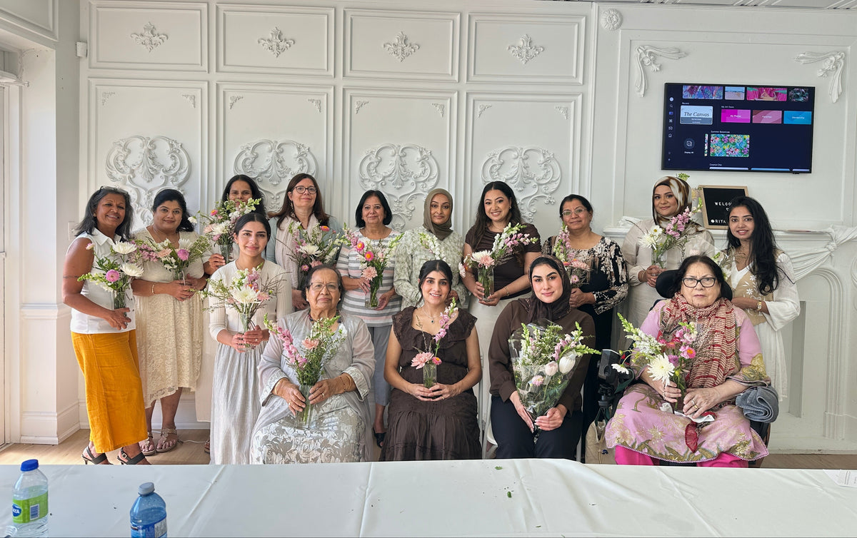 A group of women posing together in an elegant white room with decorative wall panels, holding bouquets of flowers. Some women are wearing traditional outfits.