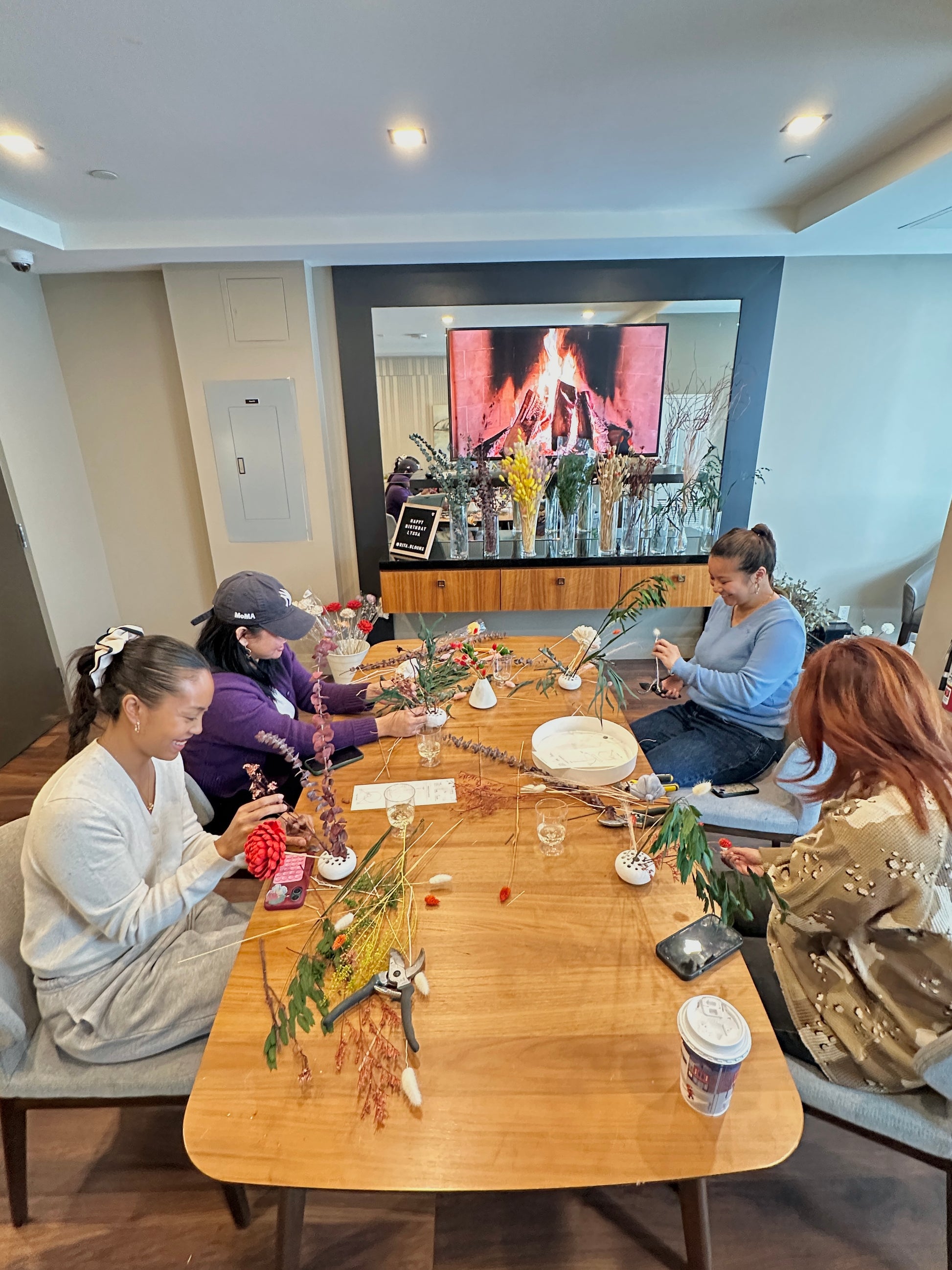 Group of people sitting around a wooden table with flowers and a screen displaying a fireplace.
