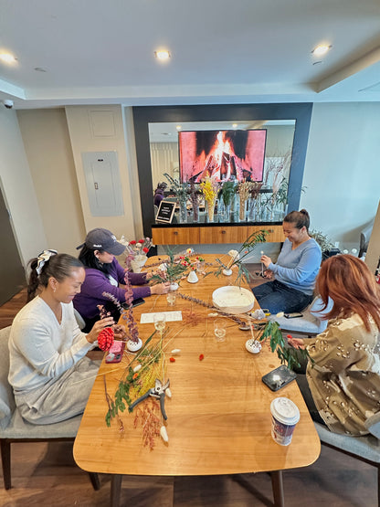 Group of people sitting around a wooden table with flowers and a screen displaying a fireplace.