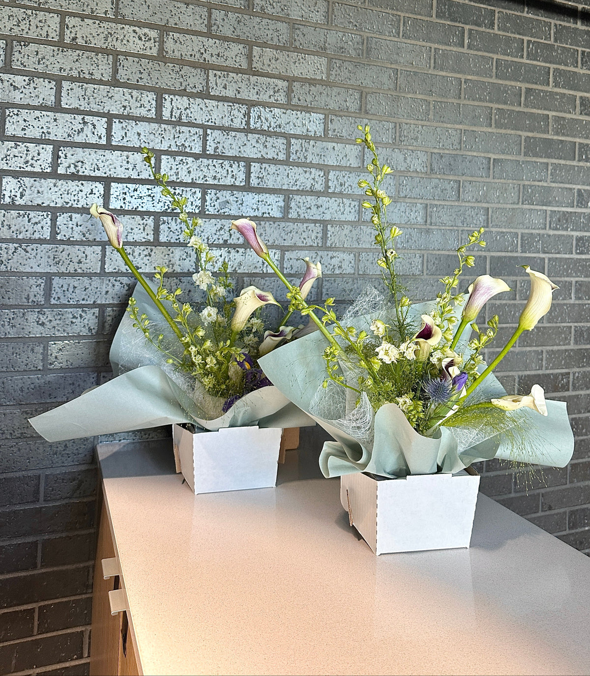 Two floral arrangements in white square pots on a light countertop, set against a gray brick wall.
