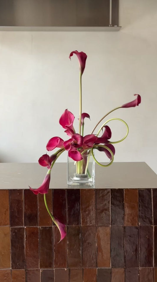 A floral arrangement of deep magenta calla lilies in a clear square glass vase, displayed on a tiled brown counter against a light wall.