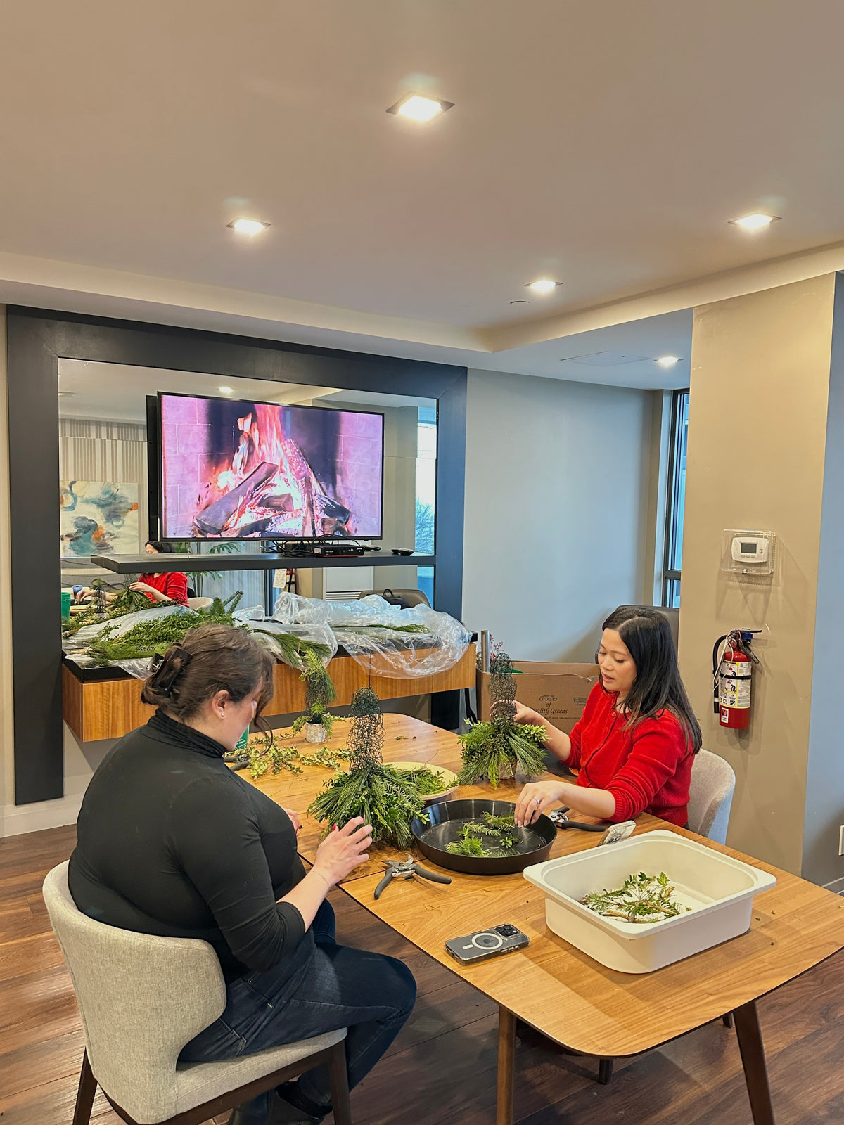 Two women are seated at a wooden dining table in a modern home interior, crafting with greenery and small plants. A large TV mounted on the wall displays a volcano scene.