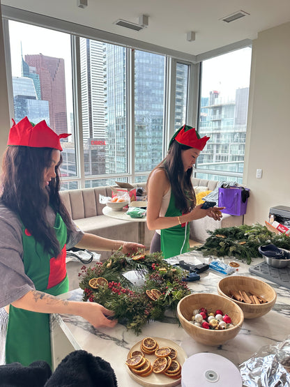 Two women in festive outfits standing near a table with holiday decorations in a room with large windows.