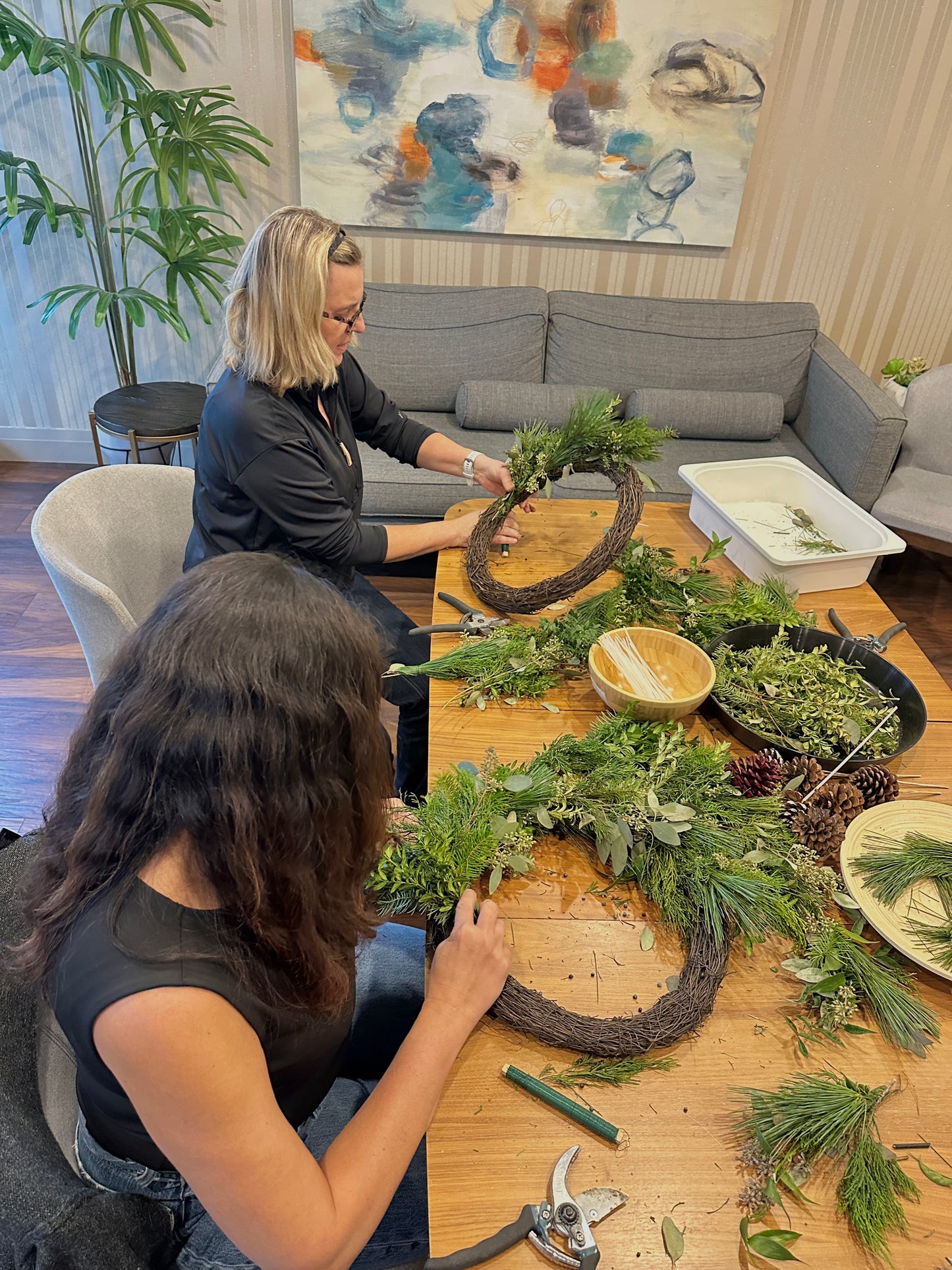 Two women crafting wreaths at a table with decorative items in a living room.