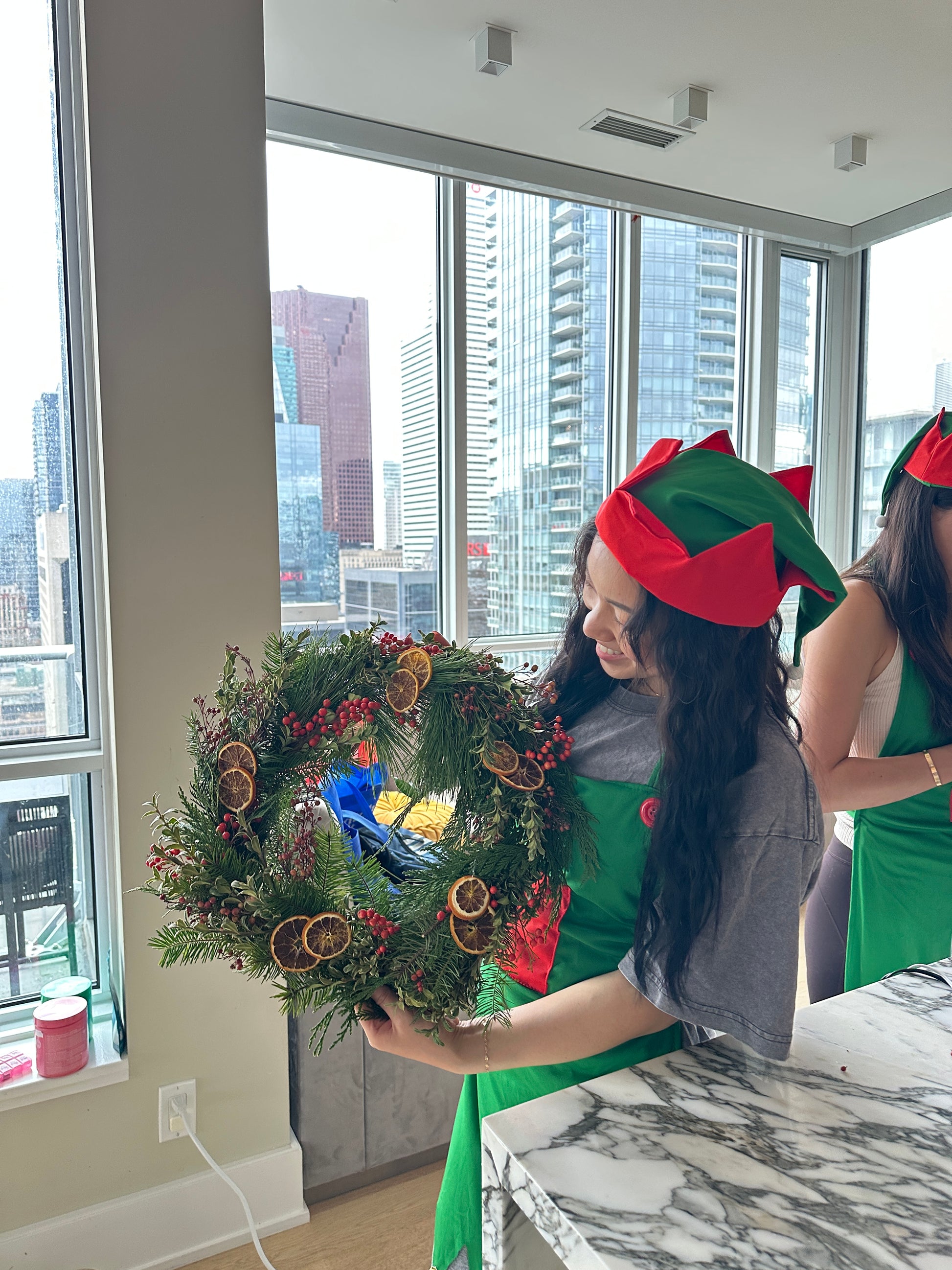 Person wearing a Christmas elf hat holding a decorative wreath indoors with a cityscape view.